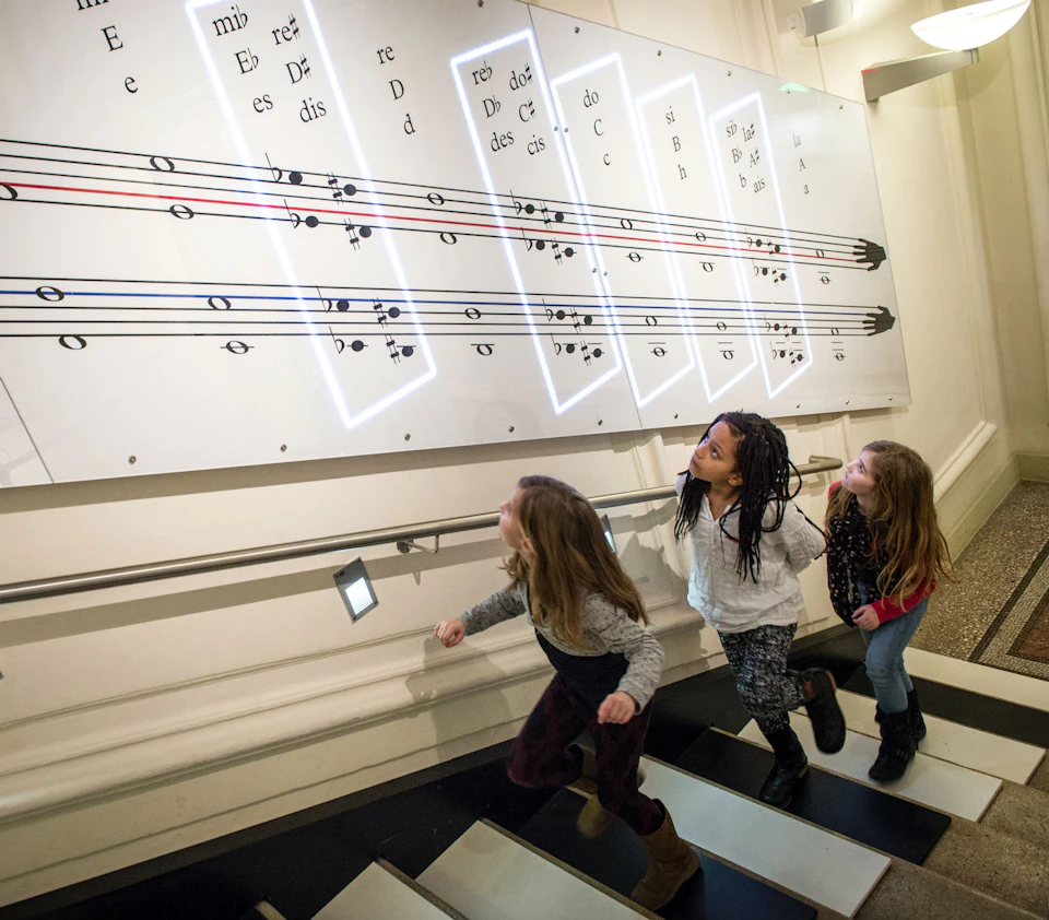 Three children ascend stairs, each step representing musical notes, while large musical notation panels are displayed on the wall.