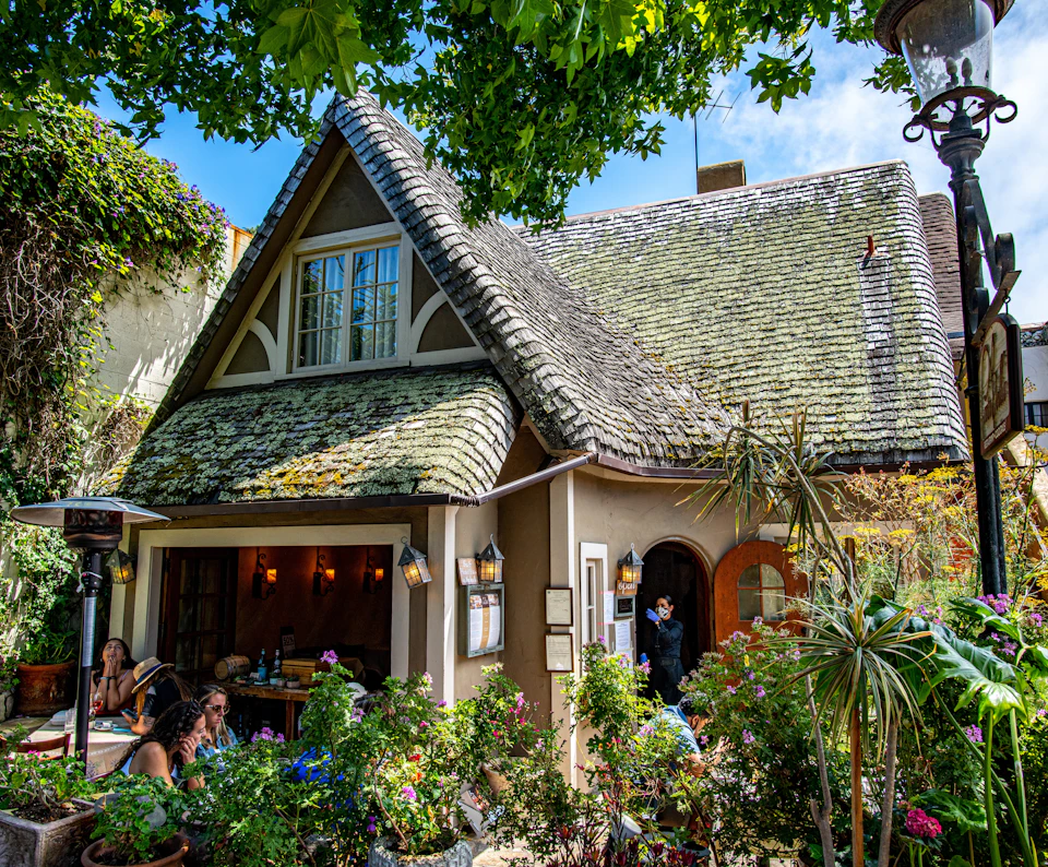 Charming cottage-style café with patrons dining outside, surrounded by lush greenery, under a clear blue sky.