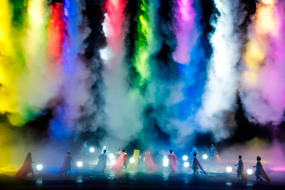 Dancers on a misty stage with colorful lights and smoke effects, holding glowing spheres, creating a vibrant atmosphere.