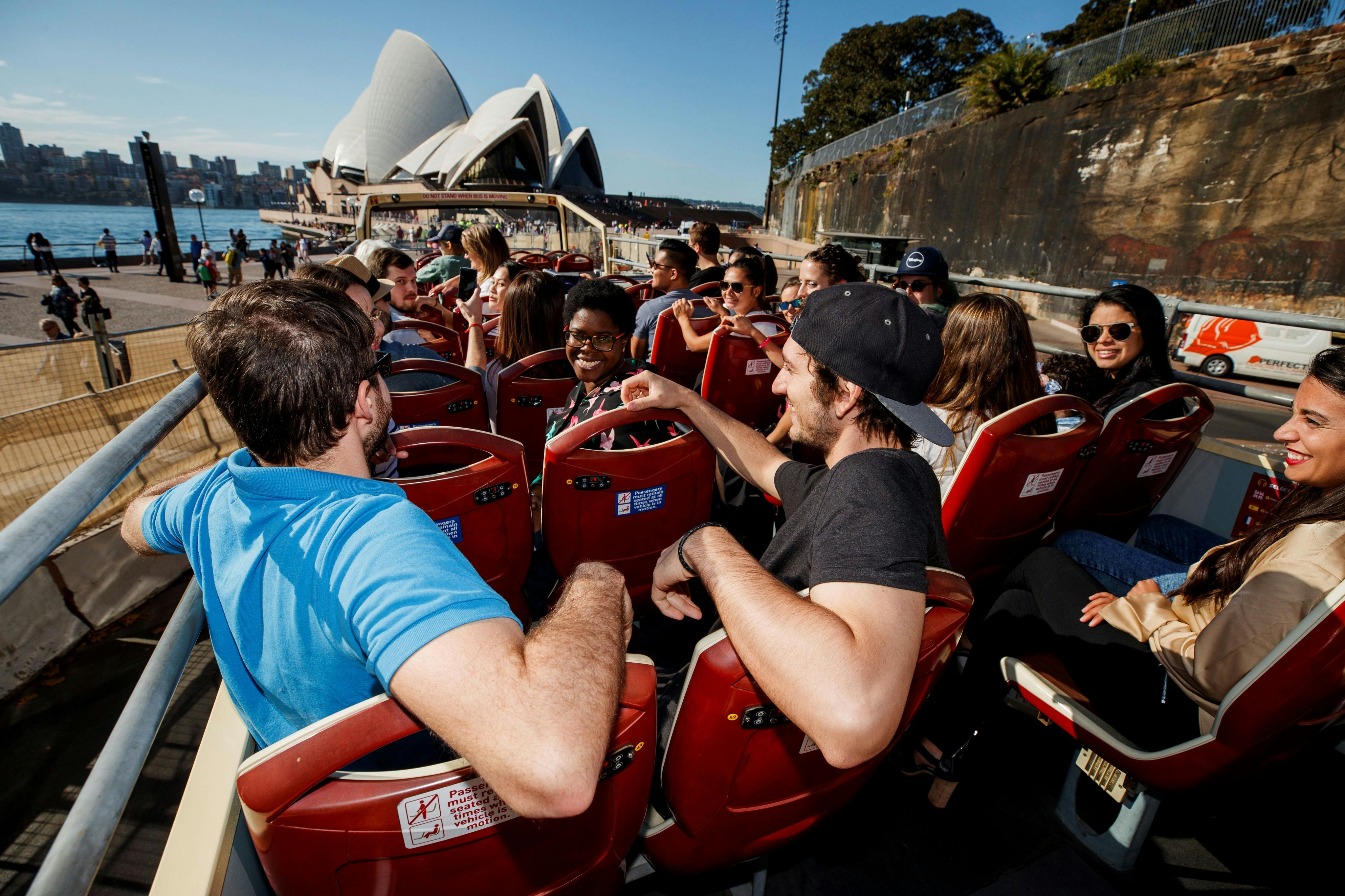 Un grup de persones que viatgen en un autobús turístic descapotable amb l'Òpera de Sydney visible al fons.
