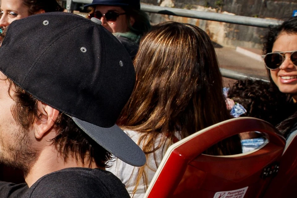 A group of people riding in an open-top tour bus with the Sydney Opera House visible in the background.