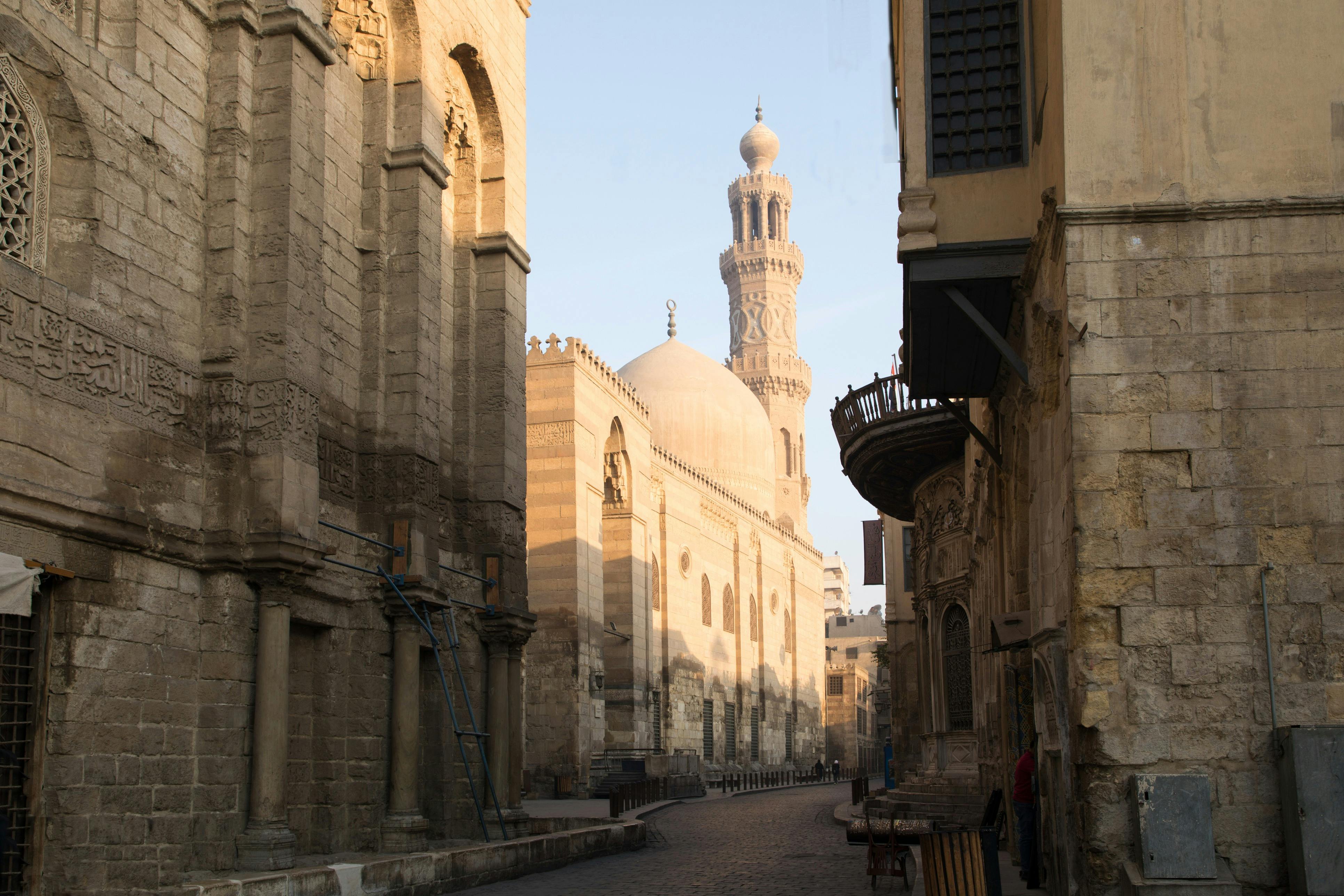 Узкая улица между историческими зданиями с выдающейся куполообразной мечетью и минаретом на заднем плане в дневное время.