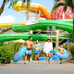 A family carrying inflatable tubes walks through a water park with colorful slides in the background.