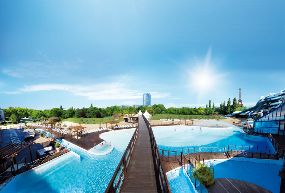 Large outdoor water park with pools, a wooden walkway, and surrounding greenery. High-rise building in the background under a bright sky.