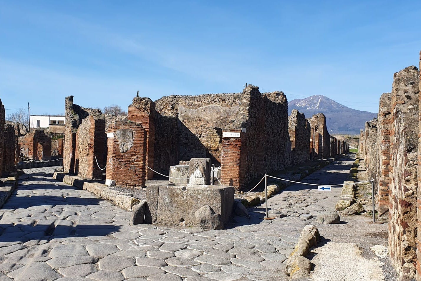 Ancient ruins with brick and stone structures, cobbled street, and a distant mountain under a clear blue sky.