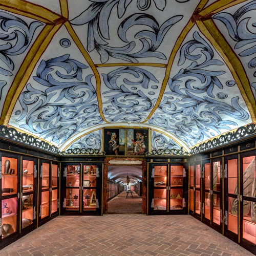 Utsmyckat rum med ett välvt, målat tak, glasskåp fyllda med artefakter och ett tegelgolv som leder till en valvgång.