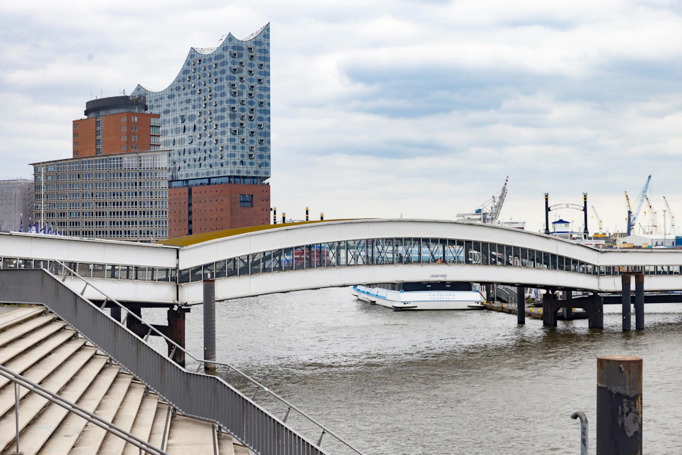 A modern building with a wavy roof, a pedestrian bridge over a river, stairs, and a docked boat under a cloudy sky.