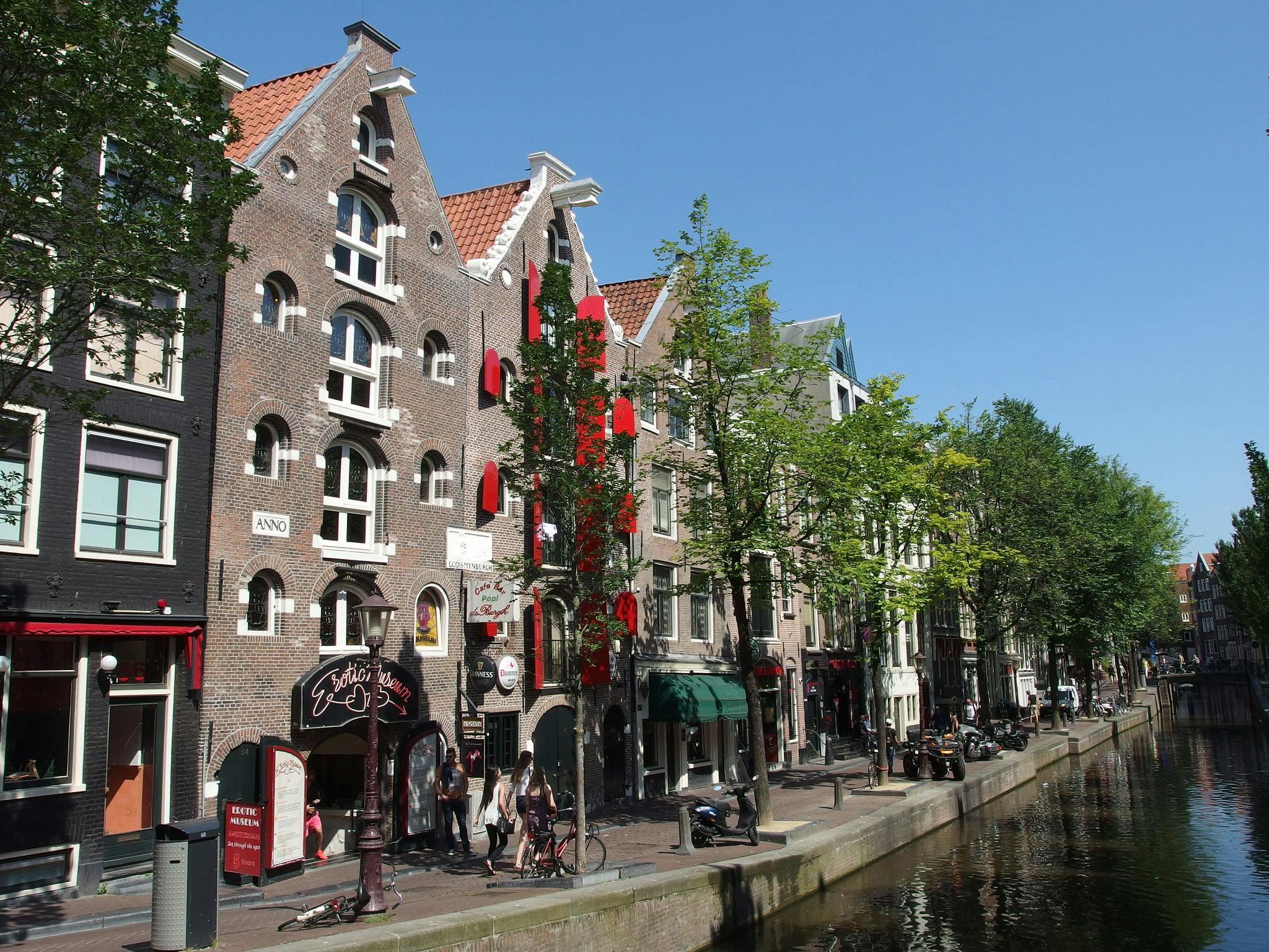 Ulica nad kanałem z zabytkowymi ceglanymi budynkami, znakami sklepowymi, drzewami i rowerami. Ludzie idą promenadą pod czystym, błękitnym niebem.