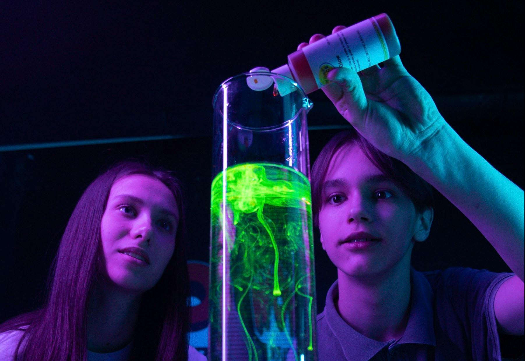 Two people observe a cylindrical container with glowing green liquid as a hand pours a substance into it.