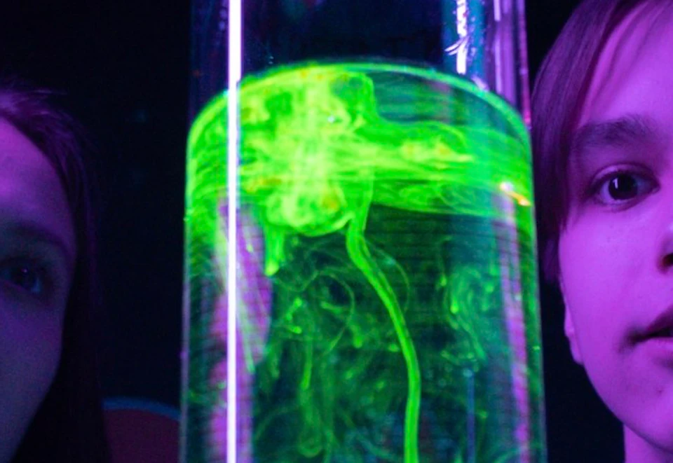 Two young individuals observe a glowing green liquid in a tall cylinder as a person pours a substance into it.