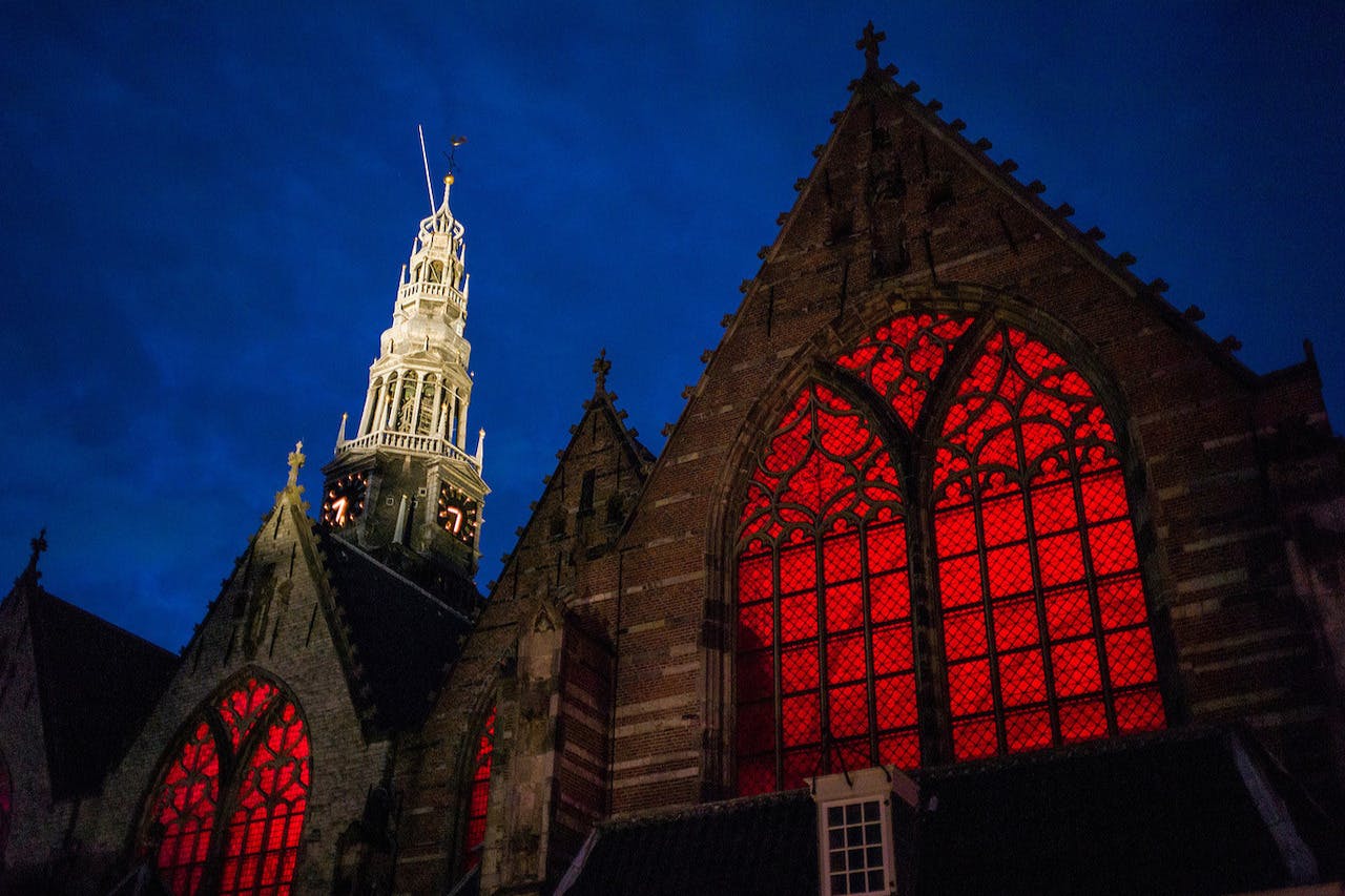 Kyrka i gotisk stil med upplysta rödmålade glasfönster och ett högt, upplyst klocktorn mot en mörkblå kvällshimmel.
