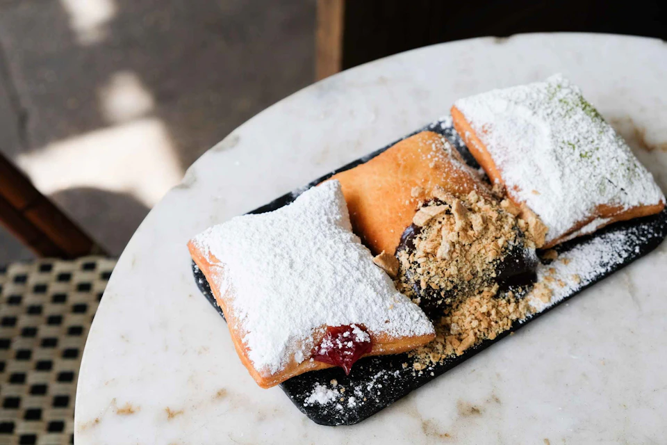 Three powdered sugar beignets, one filled with jam, on a dark rectangular plate with a chocolate and crumb topping, on a white table.