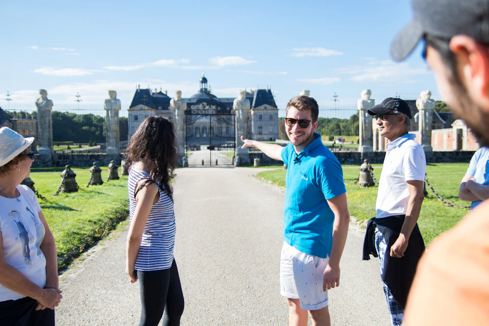 A group of people outdoors, with one person pointing towards a building in the background on a sunny day.