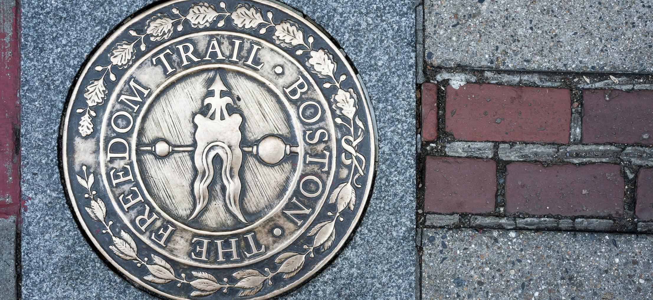 Bronze circular plaque embedded in the sidewalk, reading "The Boston Trail," adorned with leaf patterns and heraldic symbols.