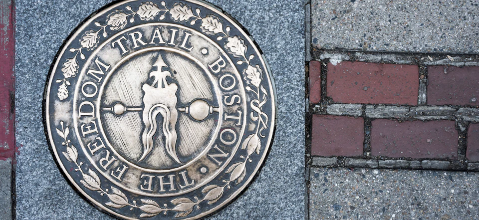 Bronze circular plaque embedded in the sidewalk, reading "The Boston Trail," adorned with leaf patterns and heraldic symbols.