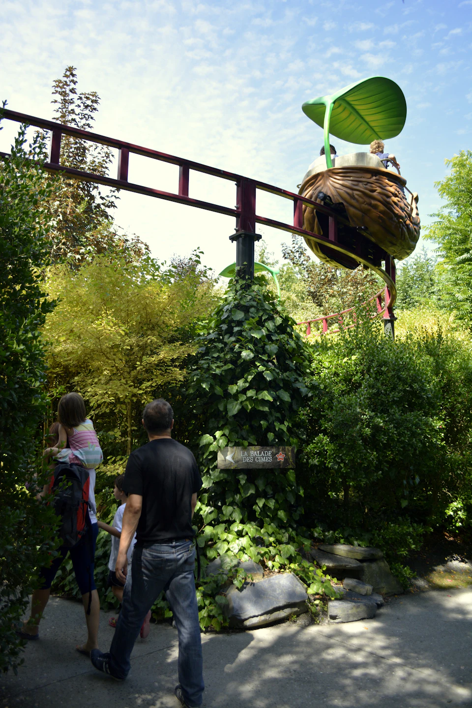 A family walks under a red roller coaster track, surrounded by dense greenery and trees, with a nest-themed ride cart above.