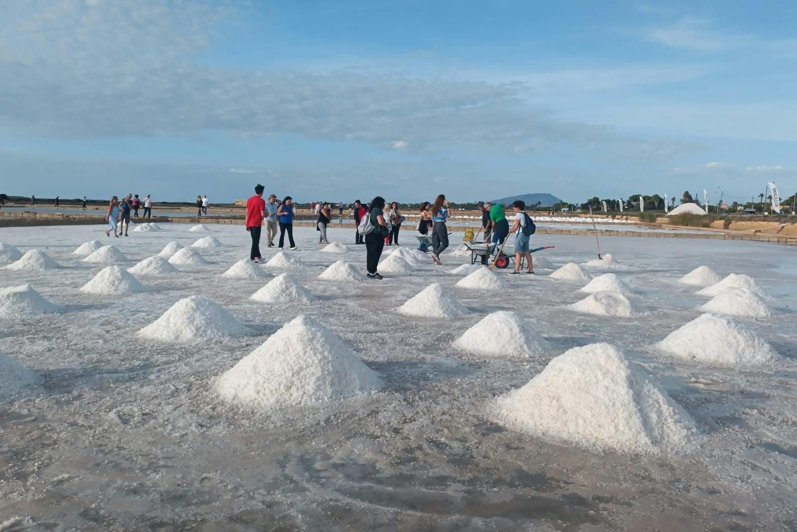 People walking and working among piles of white salt on a flat plain under a blue sky, with distant greenery and hills.
