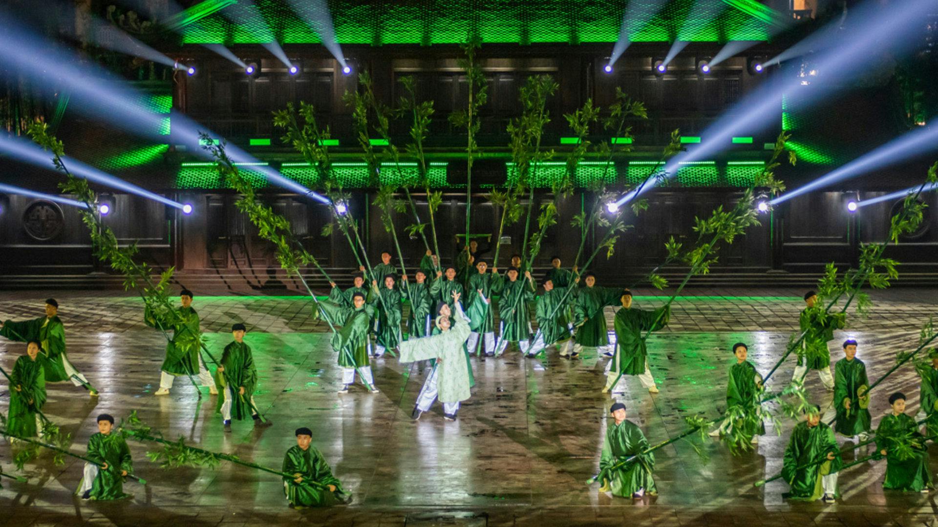 A group of performers in green costumes holding bamboo stalks, on a lit stage with green lights and spotlights.