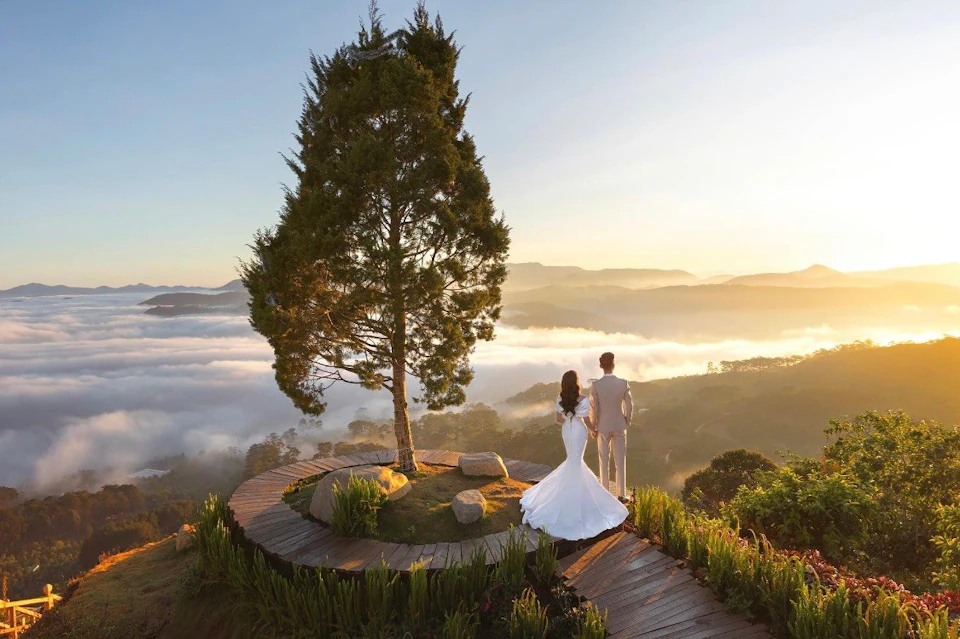 A couple in formal attire stands on a wooden platform by a lone tree, overlooking a sunrise-lit landscape with clouds and hills.