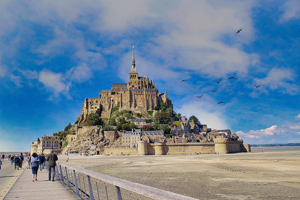 A historic castle complex on a rocky island with a spire, surrounded by medieval structures and a vast sandy foreground.