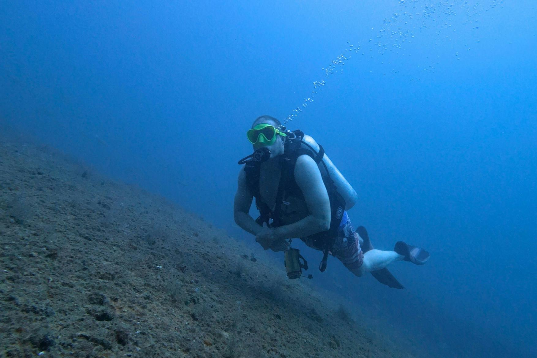 Ένας δύτης κάτω από το νερό φορώντας μια δεξαμενή και μια μάσκα, κολυμπώντας κοντά στον πυθμένα του ωκεανού με ορατές φυσαλίδες αέρα να ανεβαίνουν.