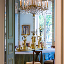 Elegant matsal med en utsmyckad ljuskrona, ett stort runt bord utsmyckat med guldkandelabrar och ett porträtt på väggen.