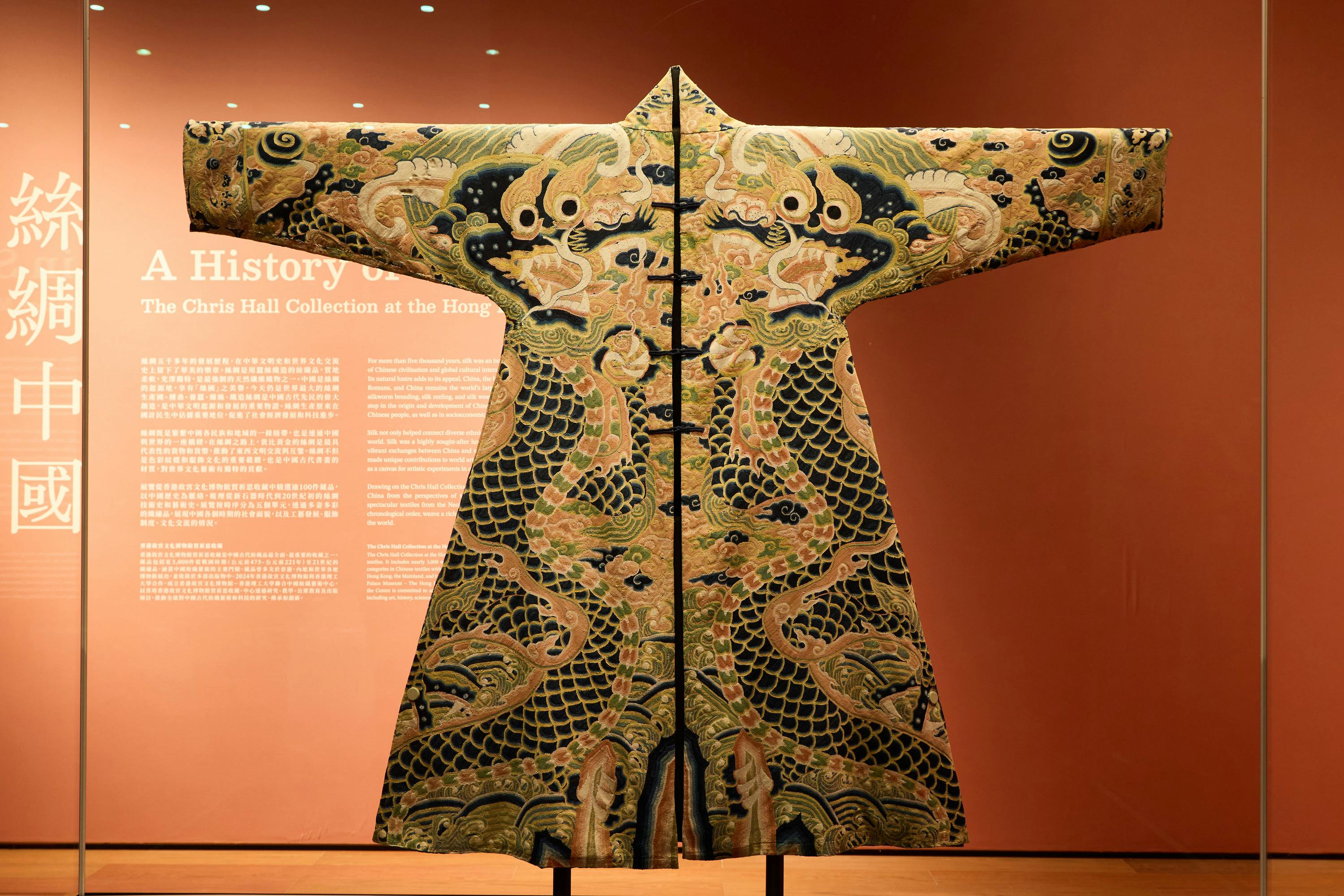 An ornate traditional robe with detailed dragon embroidery displayed against a red background in a museum setting.