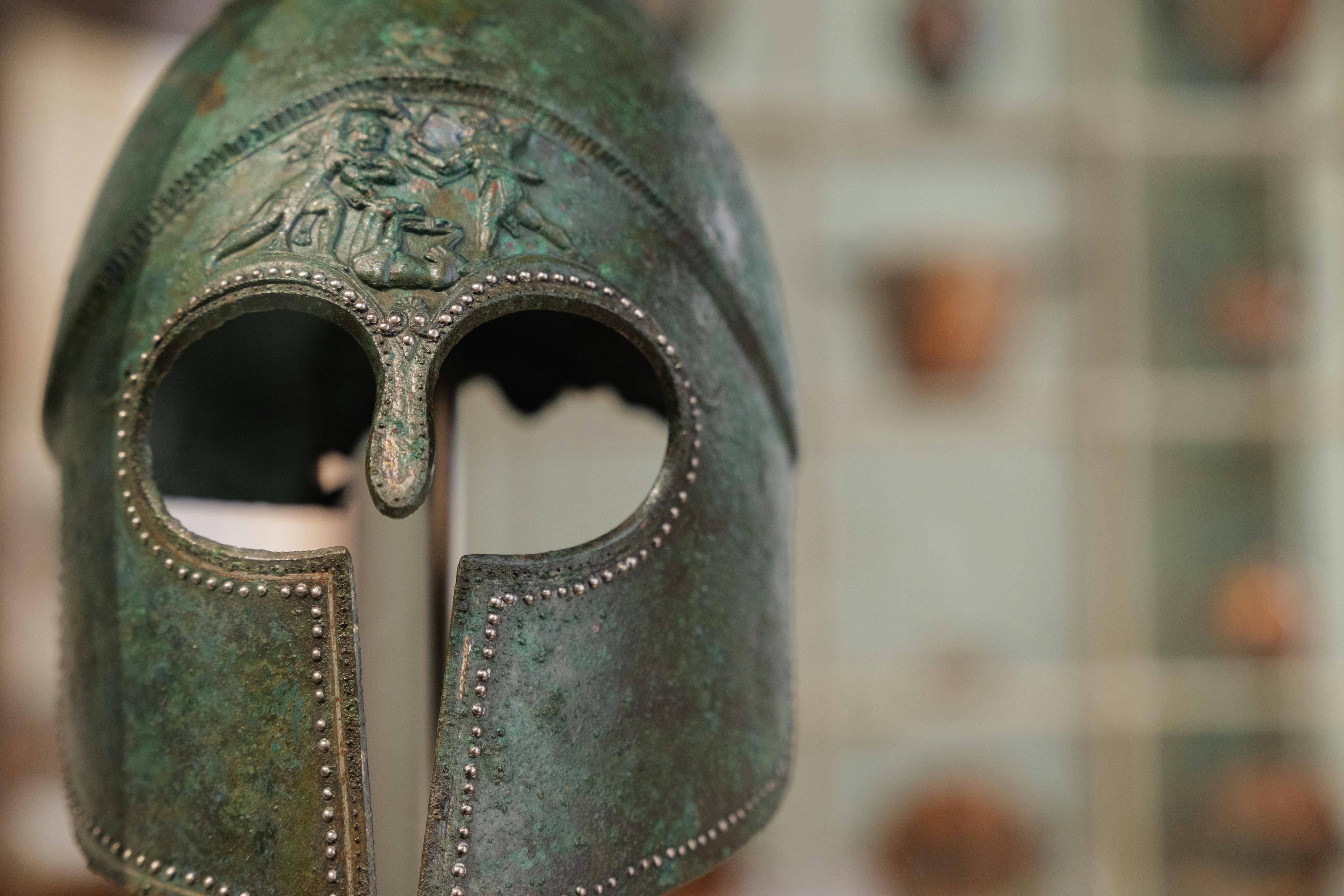 A close-up of an ancient bronze helmet displayed in a museum, with intricate detailing and a blurred background.