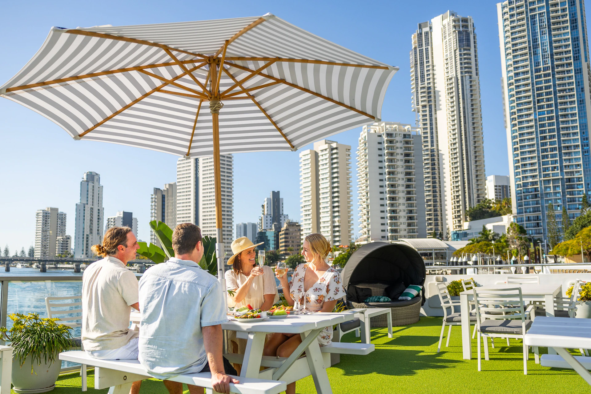 Four people sit at an outdoor table under a large striped umbrella, with tall city buildings in the background.