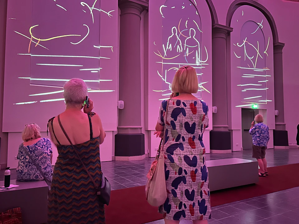 People in a gallery with purple lighting, viewing abstract white line art projected on large wall panels.
