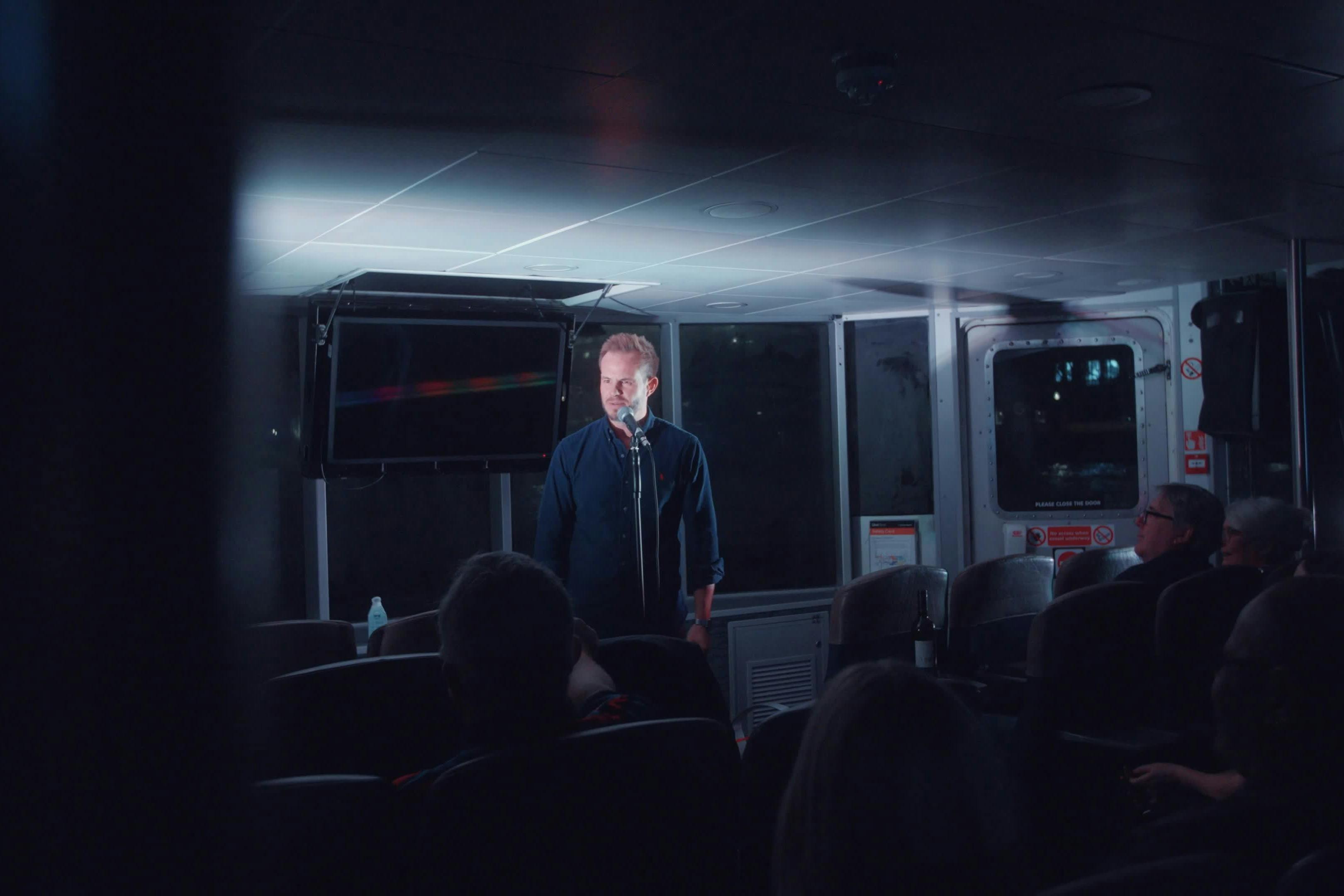 A man stands on a stage with a microphone in a dimly lit room in front of an audience.