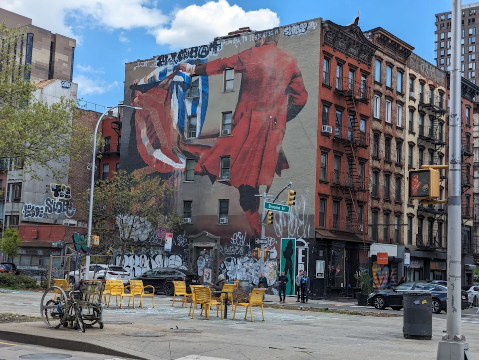 A city street corner with a large mural of a man in a red coat, outdoor seating, and graffiti-covered buildings.