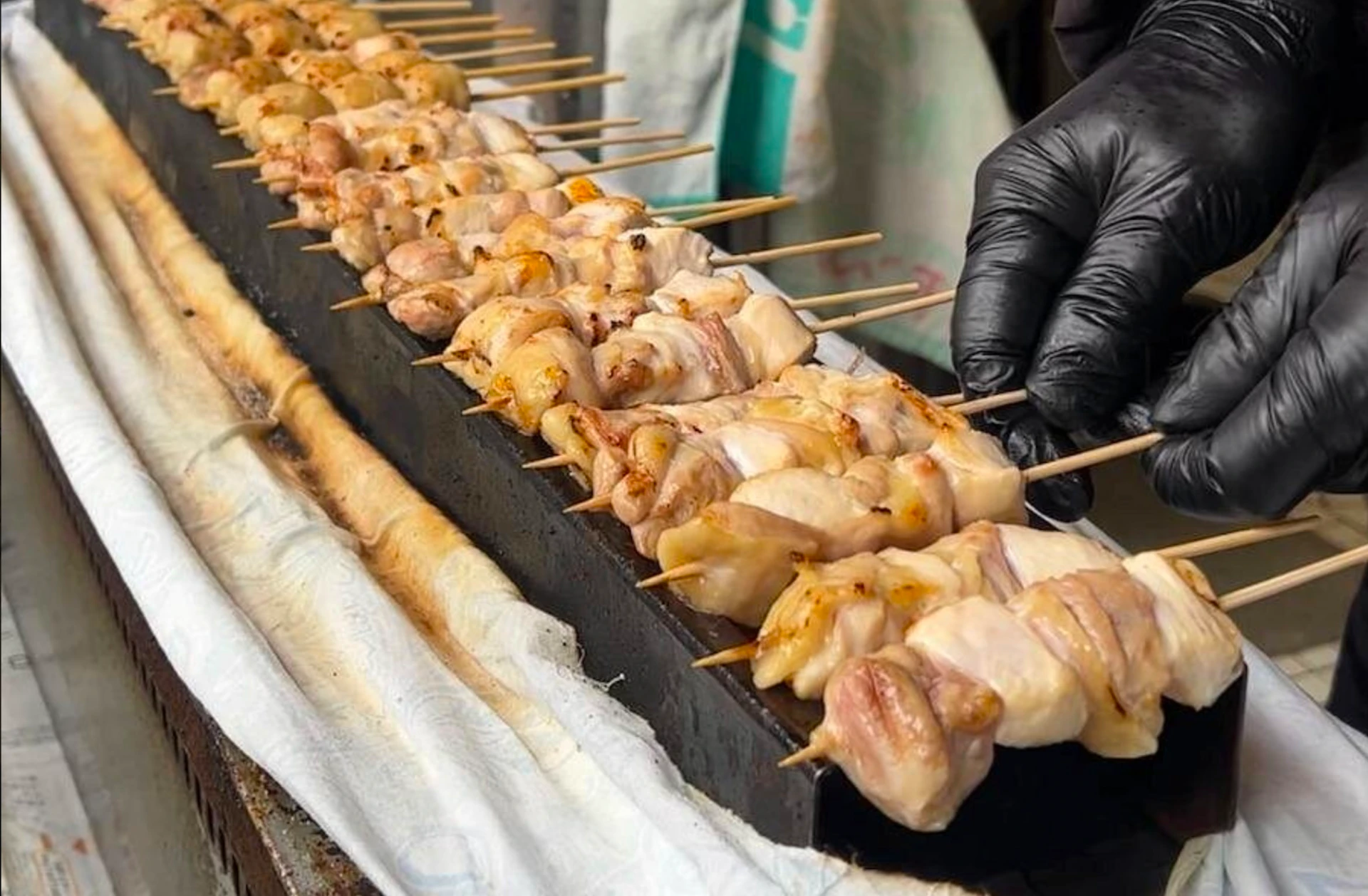 A person wearing a black glove adjusts skewers of grilled chicken on a rectangular grill with a white cloth underneath.