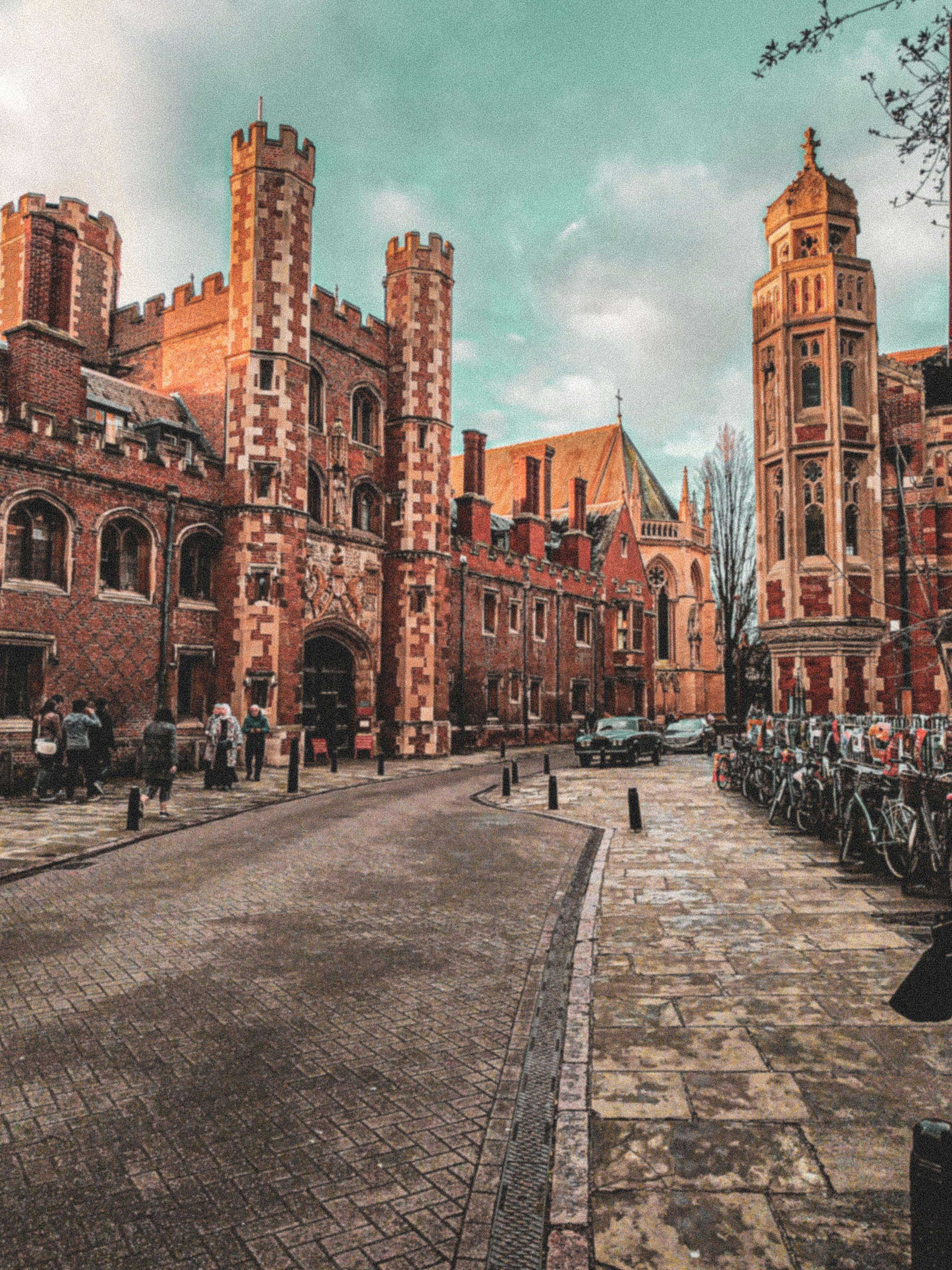 A cobblestone street lined with historic red brick buildings and bicycles, with people walking and a car parked in the background.