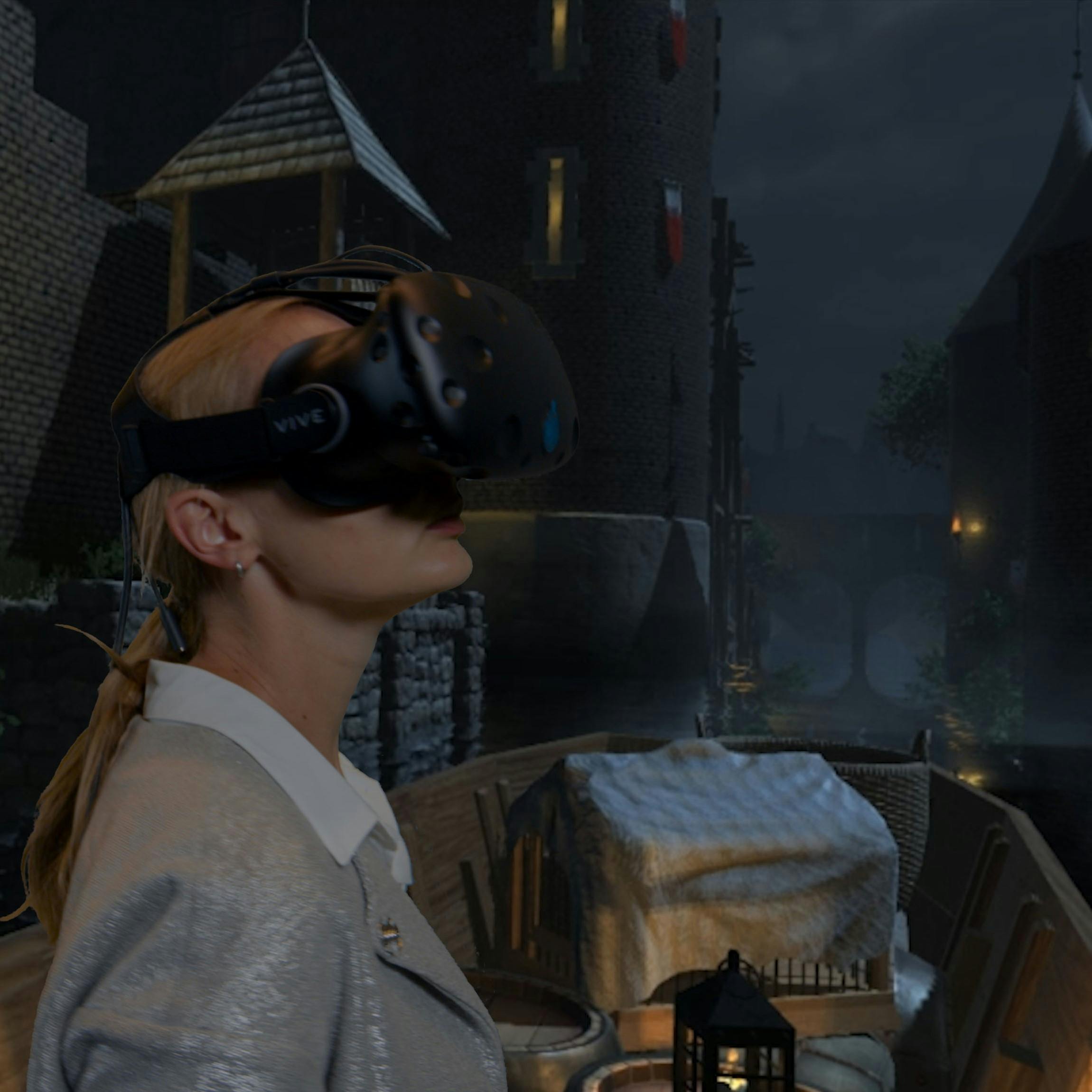 Osoba s náhlavní soupravou pro virtuální realitu ve středověkém prostředí s kamennými budovami a mostem v pozadí.