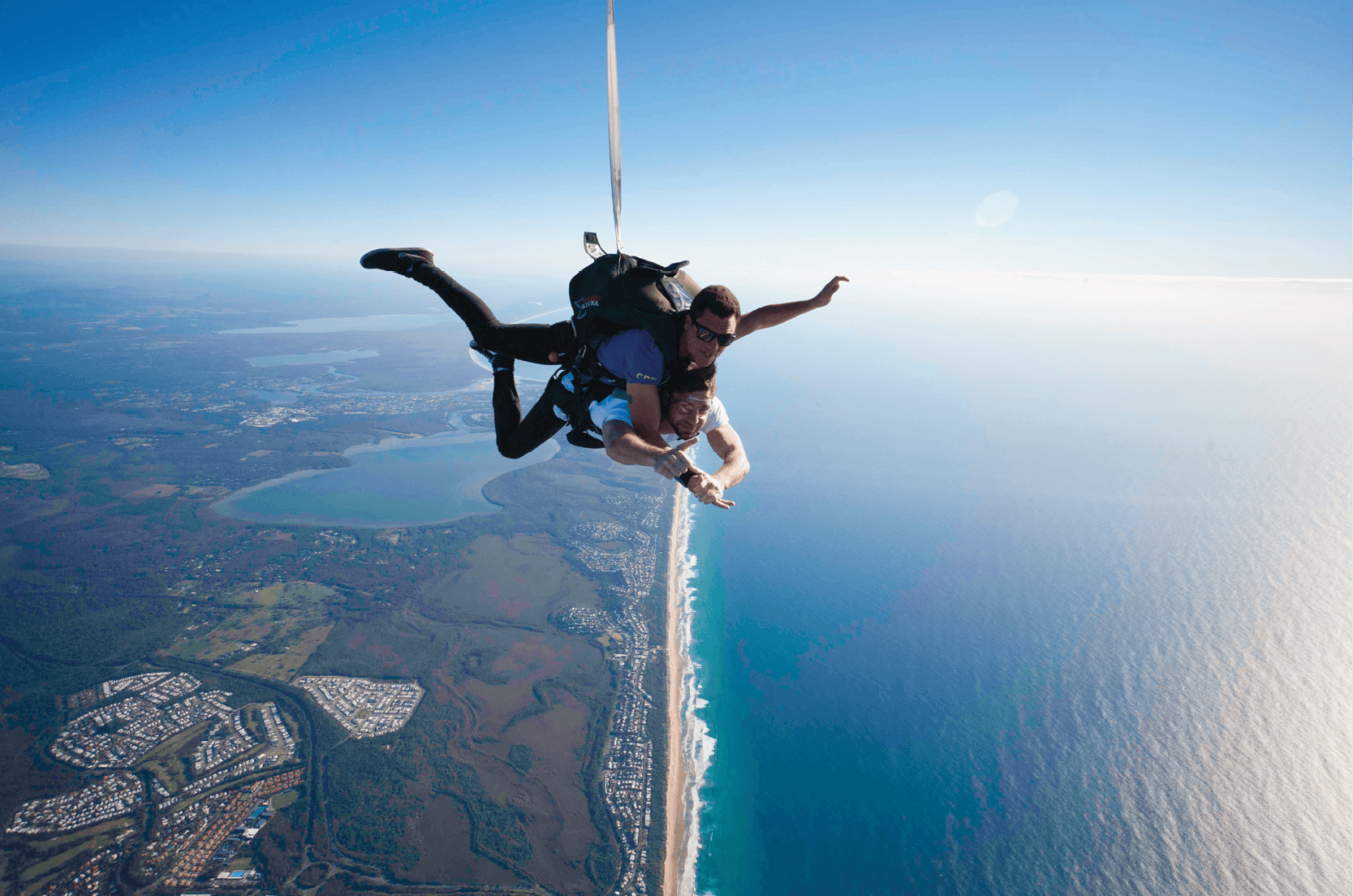 Two people are skydiving above a coastal landscape with beaches, ocean, and urban areas below.