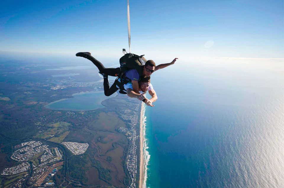Two people are skydiving above a coastal landscape with beaches, ocean, and urban areas below.