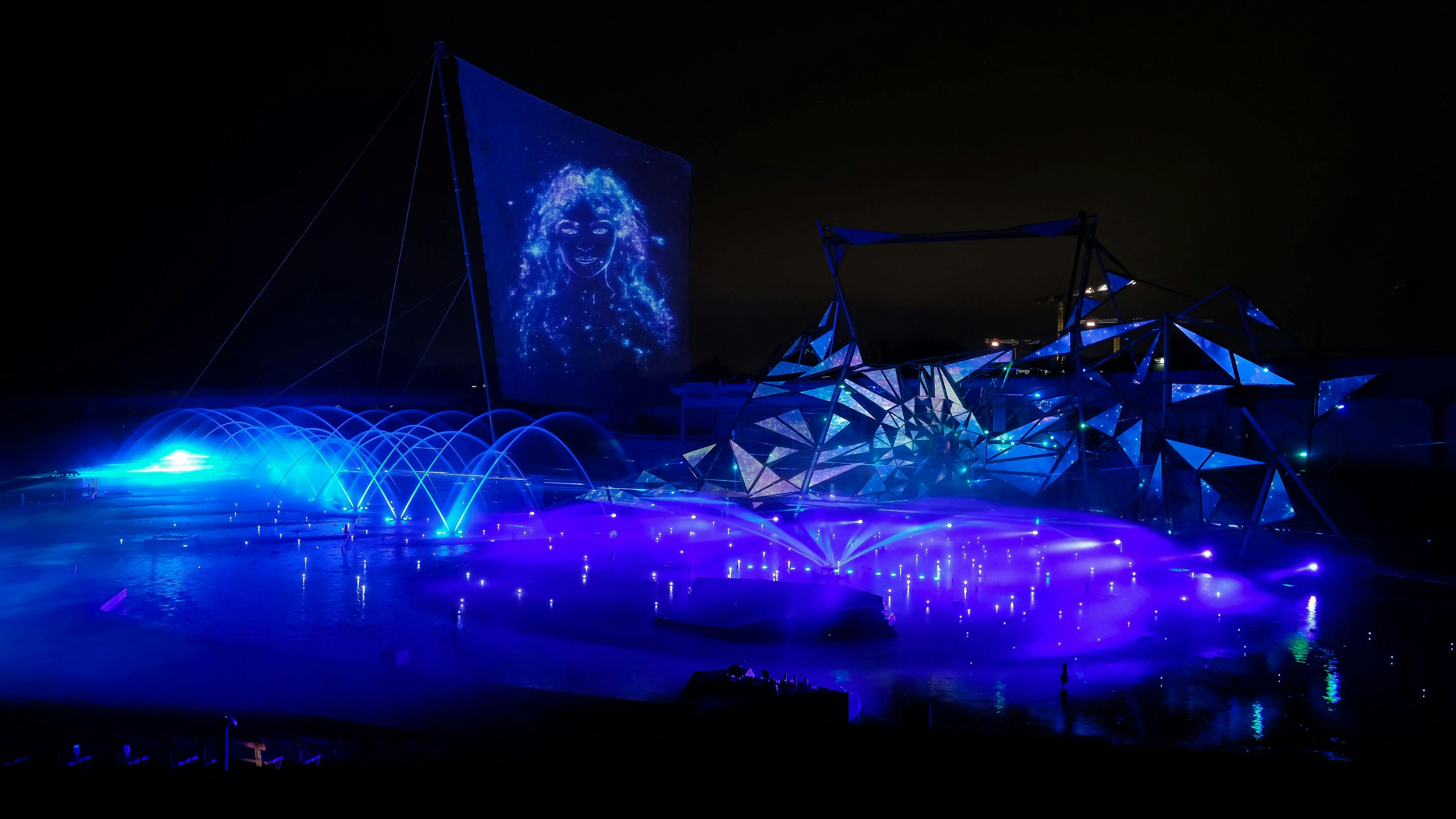 A nighttime light show with fountains, a large artistic structure, and a projected image of a face on a screen.