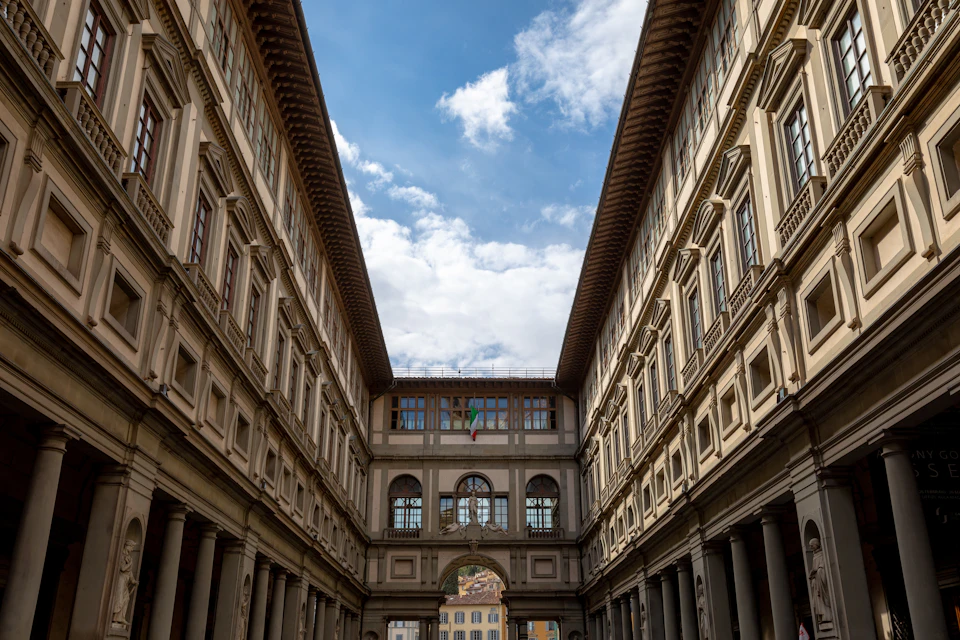 Tall, decorative stone buildings form a narrow passageway under a blue sky with scattered clouds. An archway and Italian flag are visible ahead.