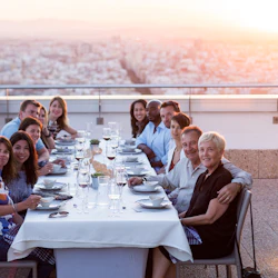 En grupp människor som sitter vid ett långt, vitklätt bord på en takterrass, med utsikt över staden i solnedgången i bakgrunden.