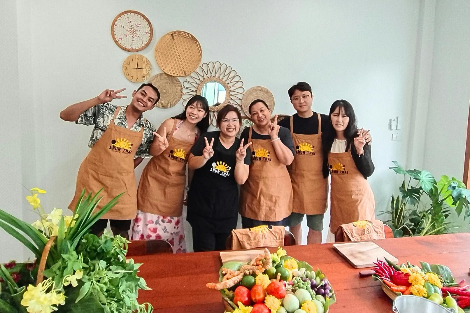 Group of six people wearing aprons posing indoors behind a table with an assortment of colorful vegetables and herbs.