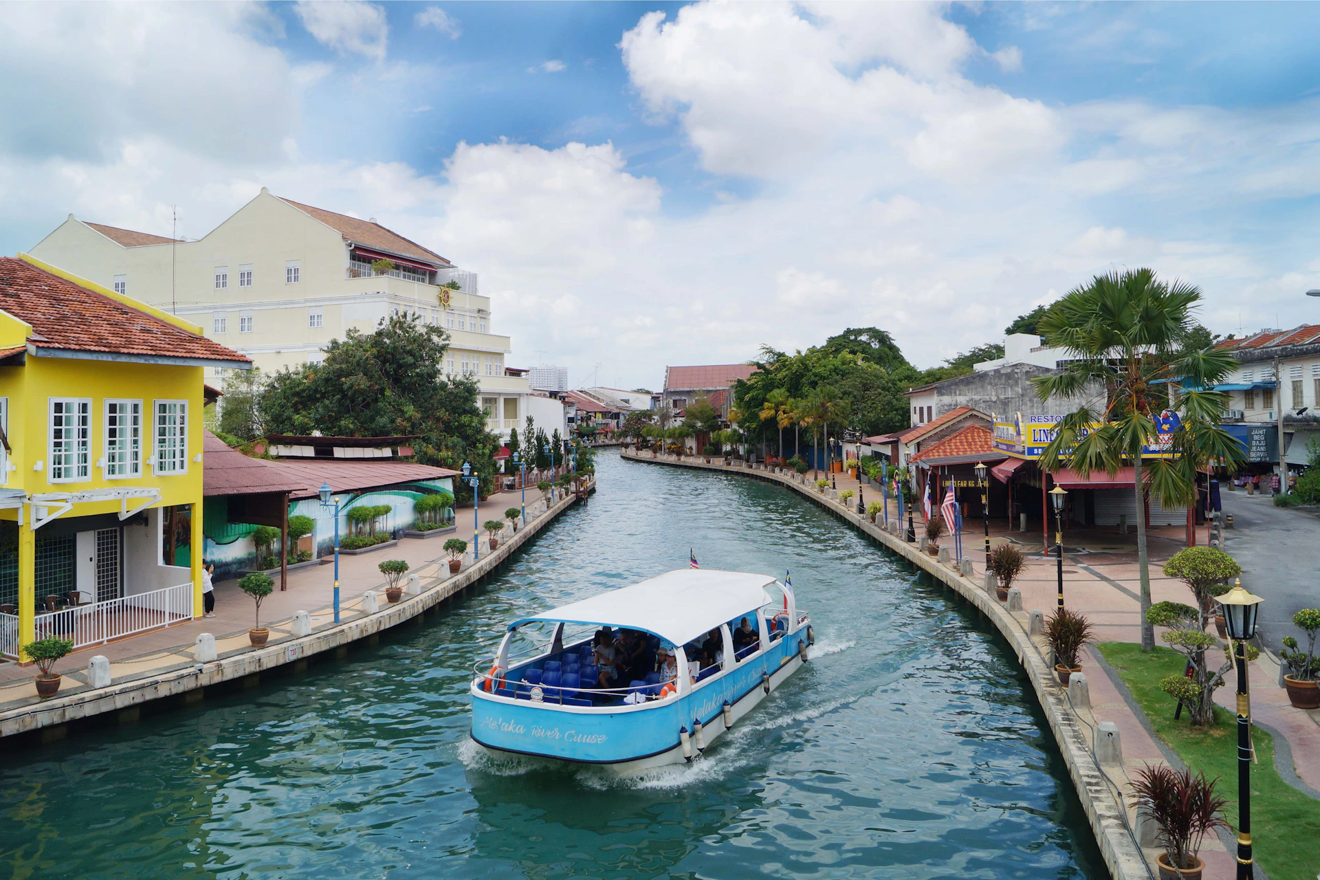 A blue and white boat cruises along a canal flanked by buildings, trees, and a pedestrian walkway under a partly cloudy sky.