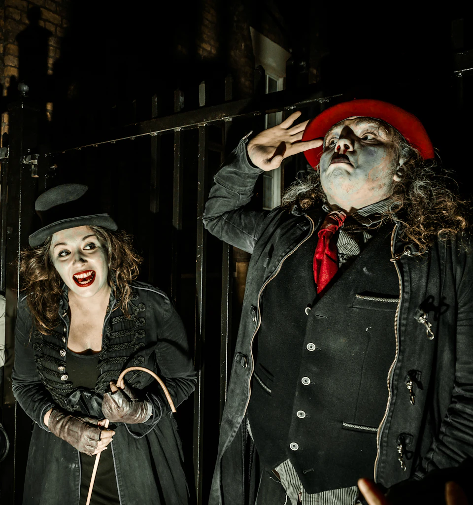 Two individuals in theatrical makeup and costumes, one with a red hat, stand next to each other against a dark, barred background.
