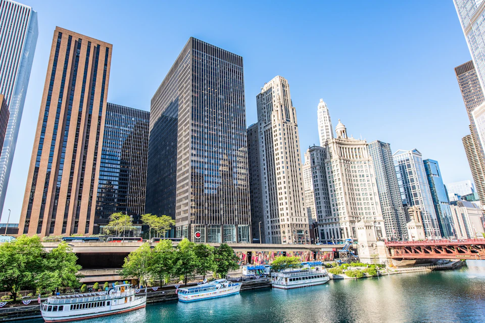 Skyscrapers along a waterfront with several boats docked below, under a clear blue sky. Trees and buildings visible in foreground.