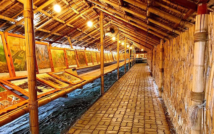 En bambusstruktur med et skråt tag viser indrammede malerier på vægge og borde, oplyst af hængende lys.