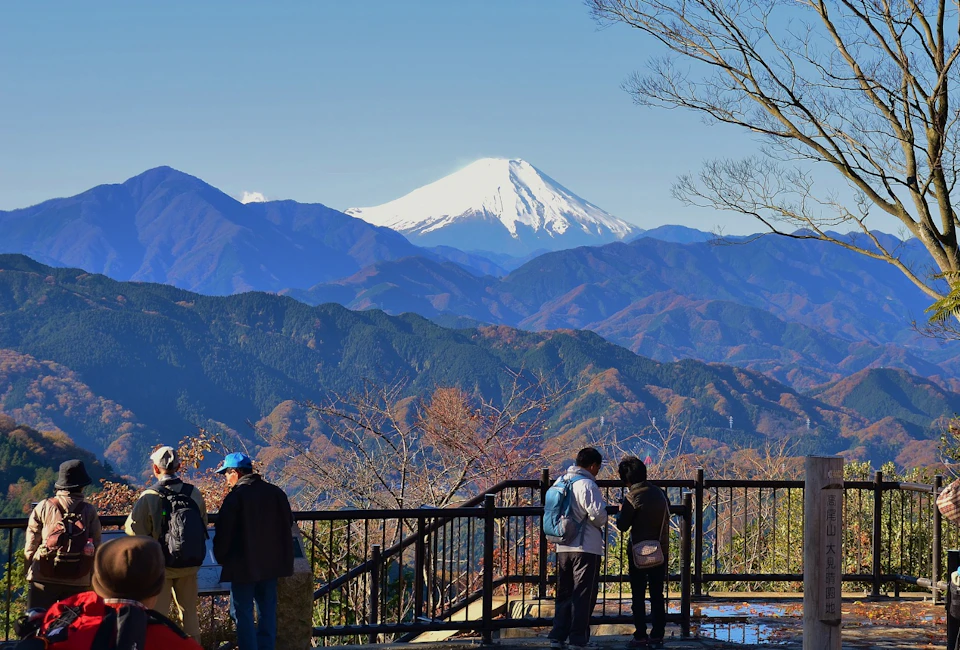 People standing on a viewing platform overlooking a mountain range with a snow-capped peak in the background on a clear day.