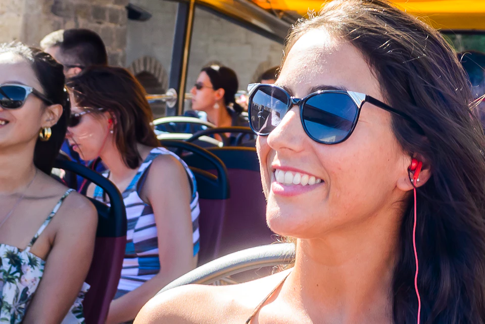 People seated on an open-top tour bus, smiling and wearing sunglasses. Historic stone arch in the background under a clear sky.