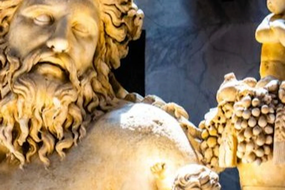 Large marble statue of a bearded man reclining, surrounded by small children. Background features additional statues and reliefs.