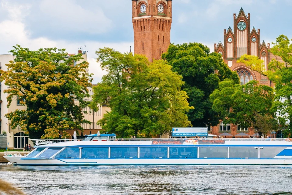 A riverboat cruising on a waterway with trees and a historic brick clock tower in the background.