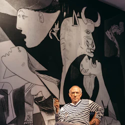 En äldre man i randig skjorta sitter på en stol och håller en cigarett med Picassos "Guernica" -målning i bakgrunden.